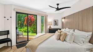 Une chambre à coucher avec un lit, un ventilateur de plafond, un banc et une vue sur un jardin, accessible par une porte coulissante.