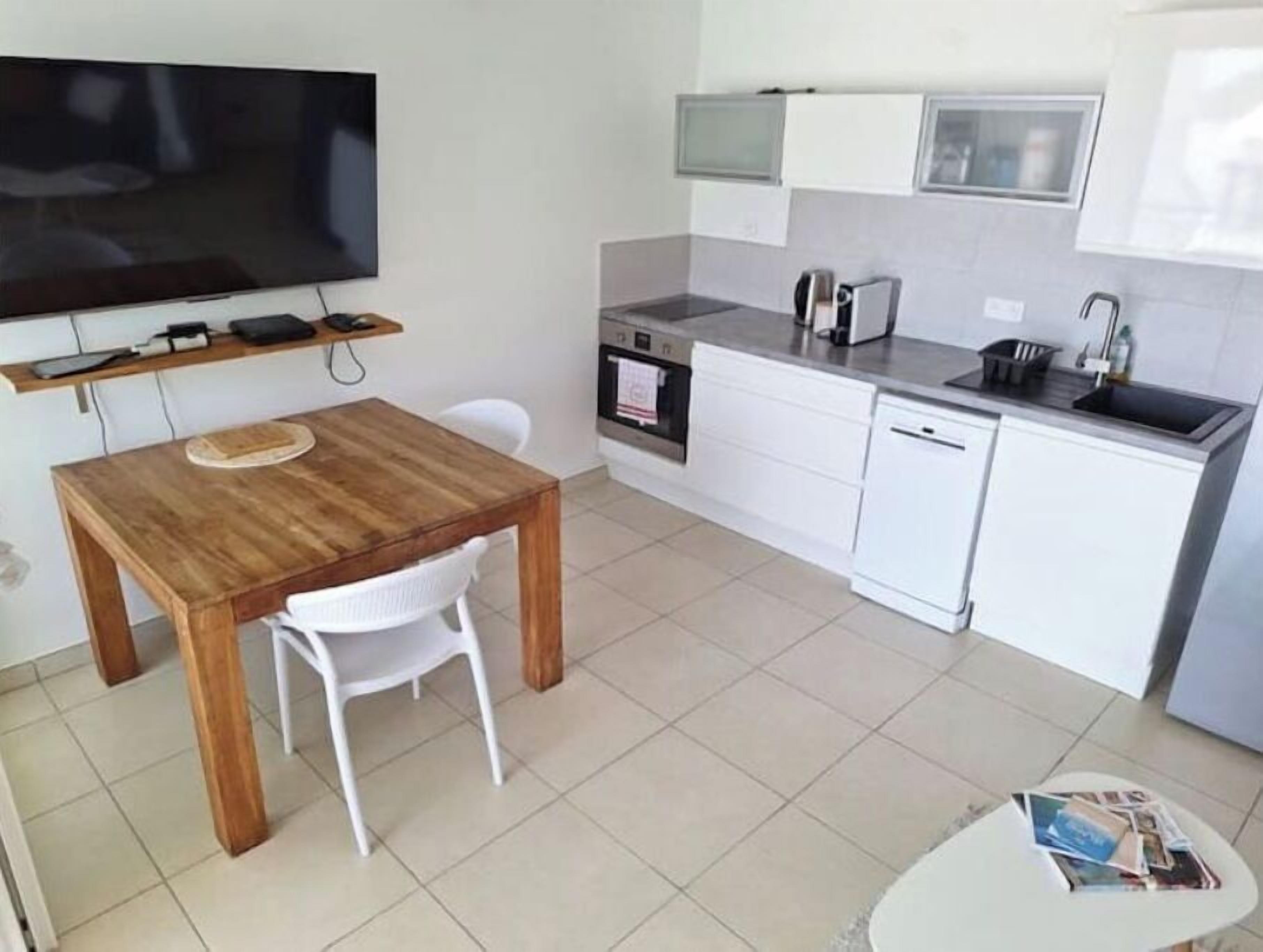 A compact kitchen with white cabinets, a wooden dining table, a white chair, a small white table, and a flat-screen TV mounted on the wall.