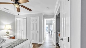 A bedroom with a bed, a ceiling fan, a nightstand with a lamp, and a hallway leading to other rooms. - Beachfront Villa W/ Pool & Tennis Hilton Head (Hilton Head Island)