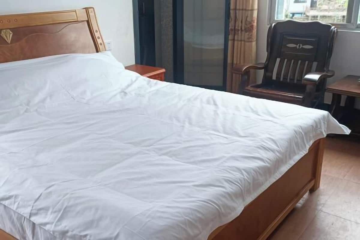 Un llit amb capçalera de fusta, una nitriera de fusta, una cadira de fusta i una finestra amb cortines.