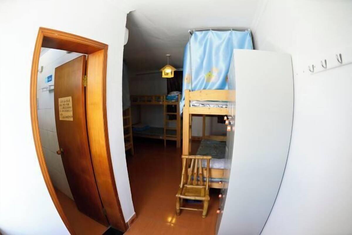 Habitació amb llits superposats, una cadira de fusta, un refrigerador blanc i una porta que dona a una altra habitació.