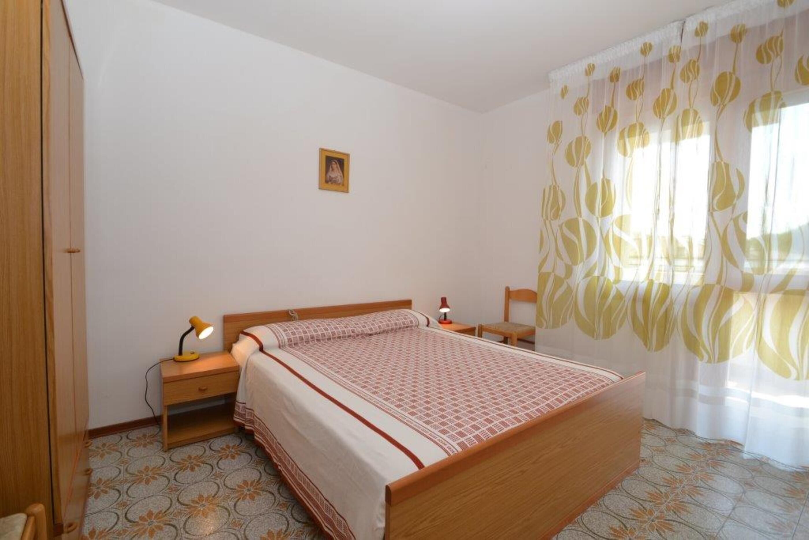 Une chambre à coucher avec un lit en bois, deux tables de chevet avec des lampes, une petite table de chevet avec une lampe, un tableau encadré au mur et une fenêtre avec des rideaux à motifs floraux.