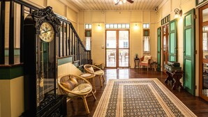 Un hall d’entrée spacieux avec un grand escalier, une grande horloge décorative, des fauteuils en osier et un tapis à motifs.