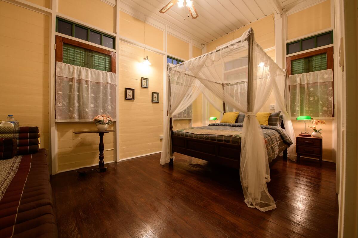 Quarto com cama de dossel, mesa de cabeceira com luminária e uma pequena mesa com um vaso de flores.