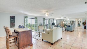 A spacious living area with a large sofa, a wooden chair, a glass-top coffee table, and a kitchen area in the background. - Oceanfront Flagler Beach Home w/ Decks & Gas Grill (Flagler Beach)