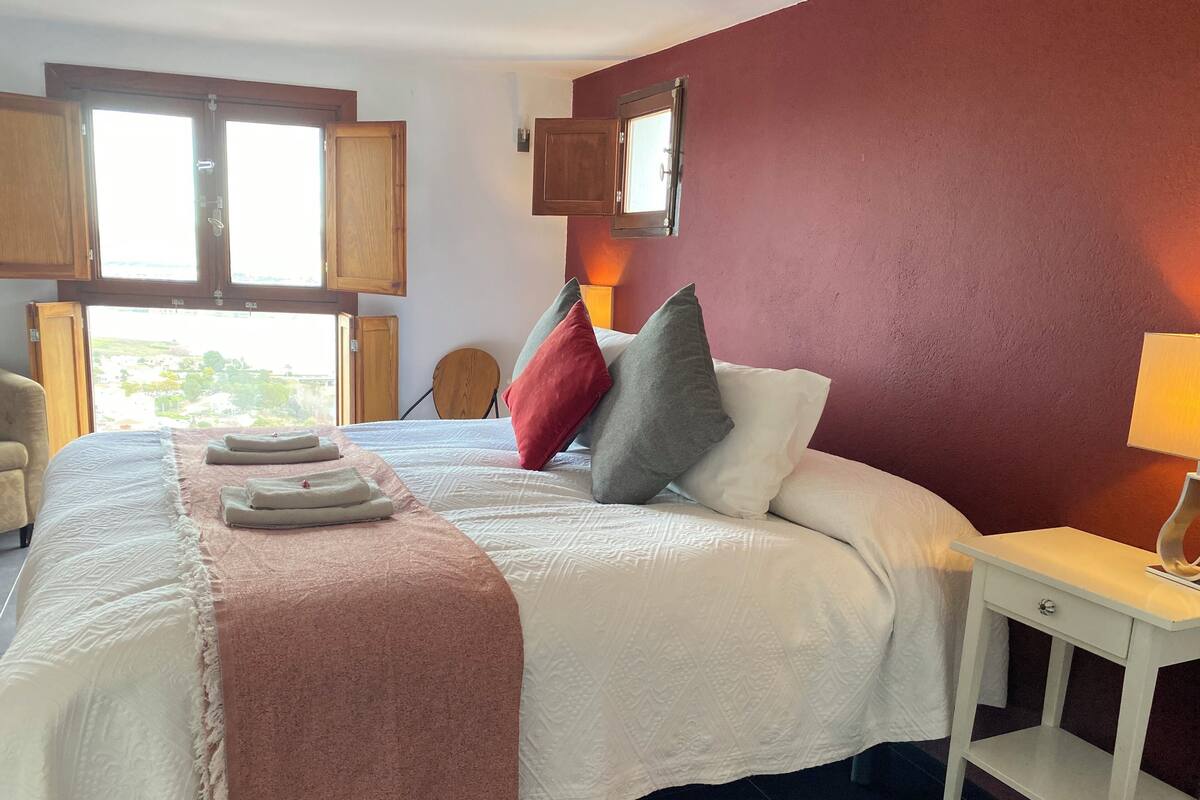 Un llit ben fet amb una manta rosa, almoinos i tovalloles netejades. Hi ha una tauleta de nit de fusta amb una làmpada i una vista a l'exterior a través d'una finestra.