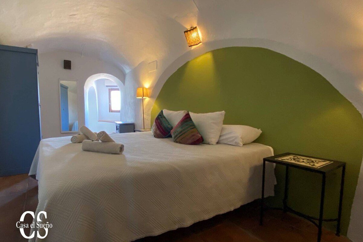 Een slaapkamer met een bed, kussens, een nachtkastje en een lamp.