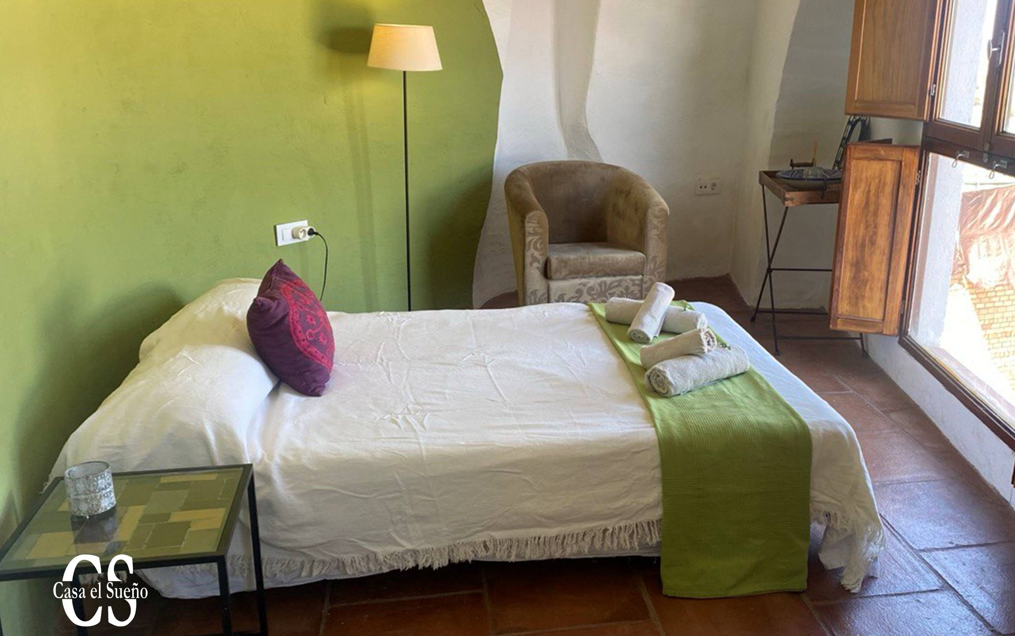 A neatly made bed with a green throw blanket, a chair, a small table with a glass container, and a lamp.
