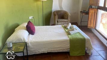 A neatly made bed with a green throw blanket, a chair, a small table with a glass container, and a lamp.