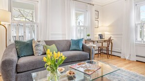 Una sala de estar con un sofá gris, una mesa de centro de vidrio y un jarrón con tulipanes amarillos. Hay un comedor con una mesa y sillas, y una ventana con cortinas blancas.