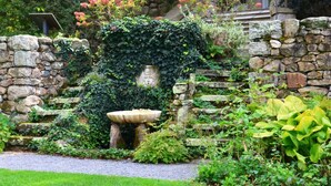 Jardín con muro de piedra, una fuente, abundante vegetación y una escalera de piedra.