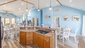 A spacious kitchen with a central island, wooden cabinets, and white chairs. - Aqua Palooza 🌊 Semi-Oceanfront 🏖️Pool & Hot Tub 💦 Elevator ⬆️ Pet Friendly🐕 (Nags Head)