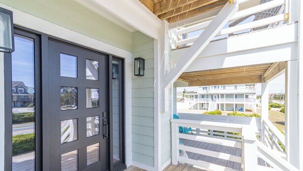 A wooden deck with a white railing, a black door with multiple windows, and a view of a building in the background. - Aqua Palooza 🌊 Semi-Oceanfront 🏖️Pool & Hot Tub 💦 Elevator ⬆️ Pet Friendly🐕 (Nags Head)