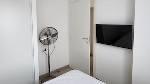 Un dormitorio con cama, ventilador de pie, televisor montado en la pared y una puerta que da a otra habitación.