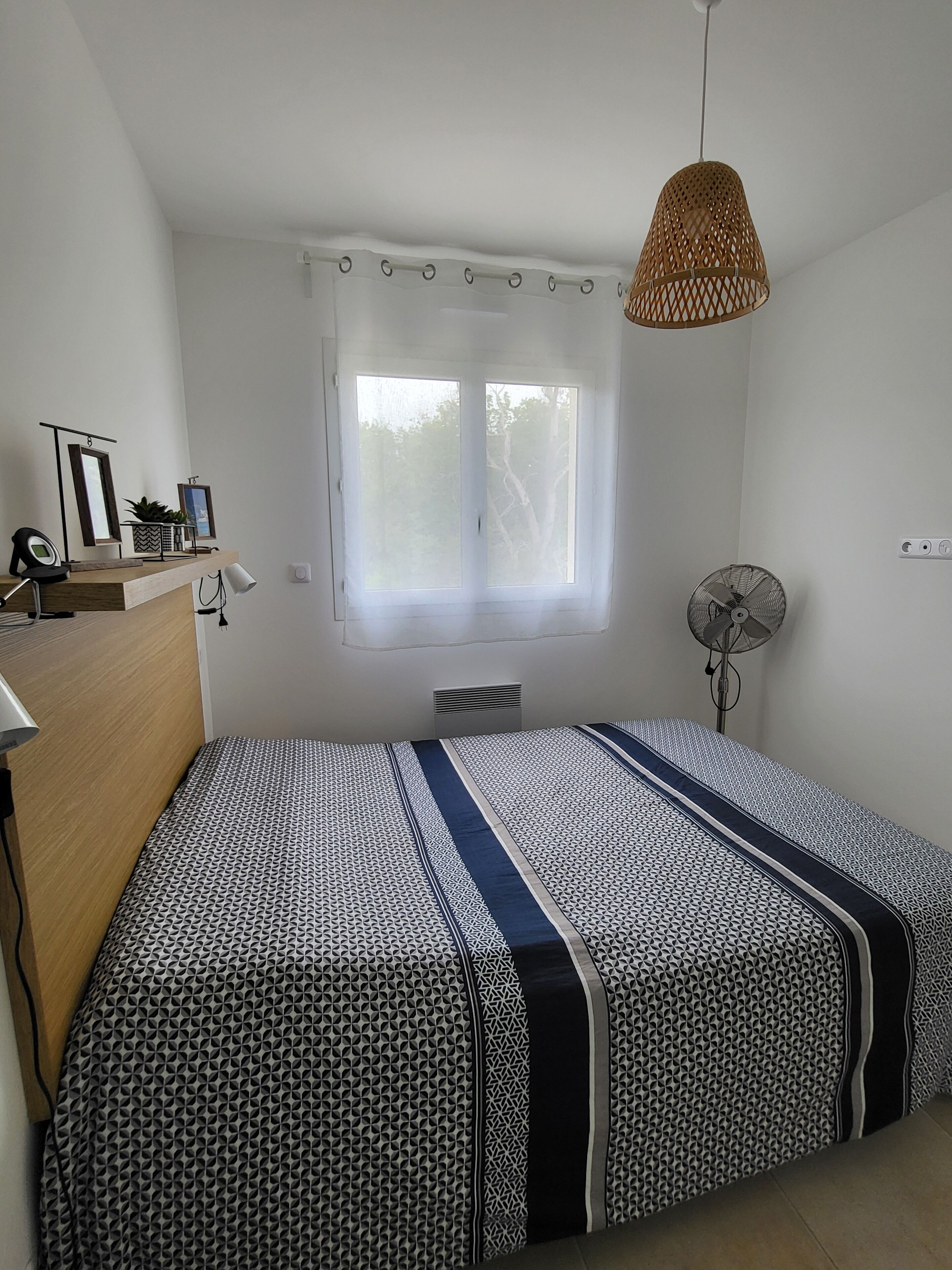 A bedroom with a bed, a window with curtains, a standing fan, and a ceiling light.