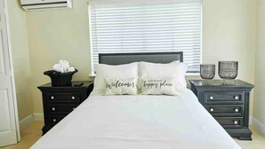 A bedroom with a bed, two bedside tables, a window with blinds, a wall-mounted air conditioner, and a nightstand with a basket and glasses. - Delva’s Place-Gated 3 bedroom - Pet Friendly (Freeport)