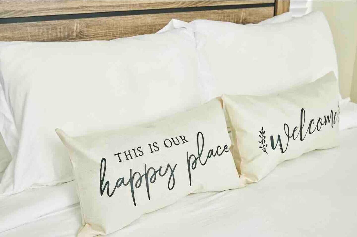Two pillows on a bed with the text "THIS IS OUR happy place" and "Welcome" on them.