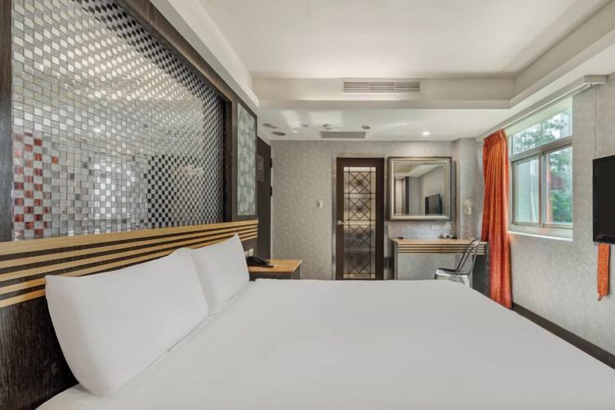 A hotel room with a large bed, a wooden headboard, a bathroom with a mirror, and a window with curtains.