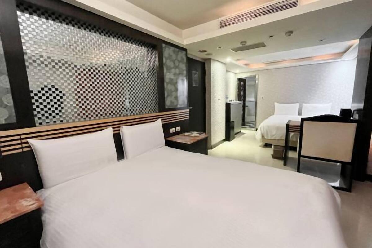 A hotel room with a large bed, a desk, and a second bed in the background.