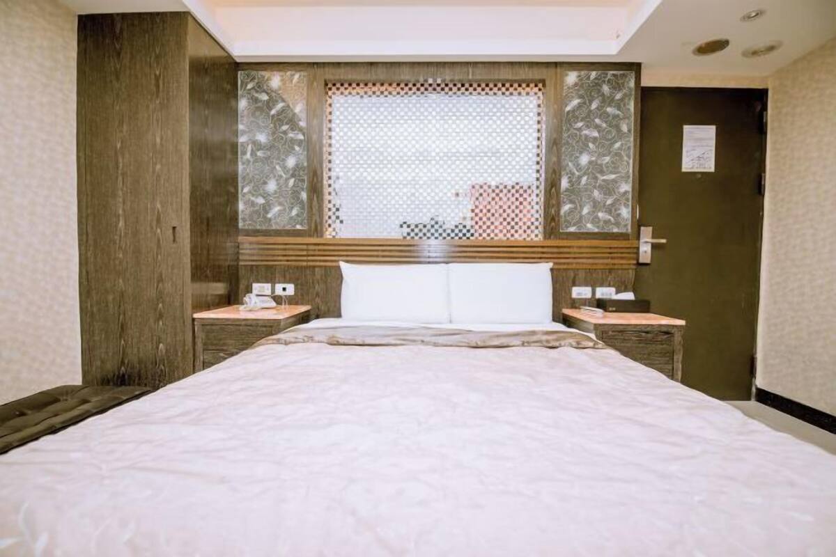 A hotel room with a bed, two bedside tables, a window with patterned curtains, and a door.