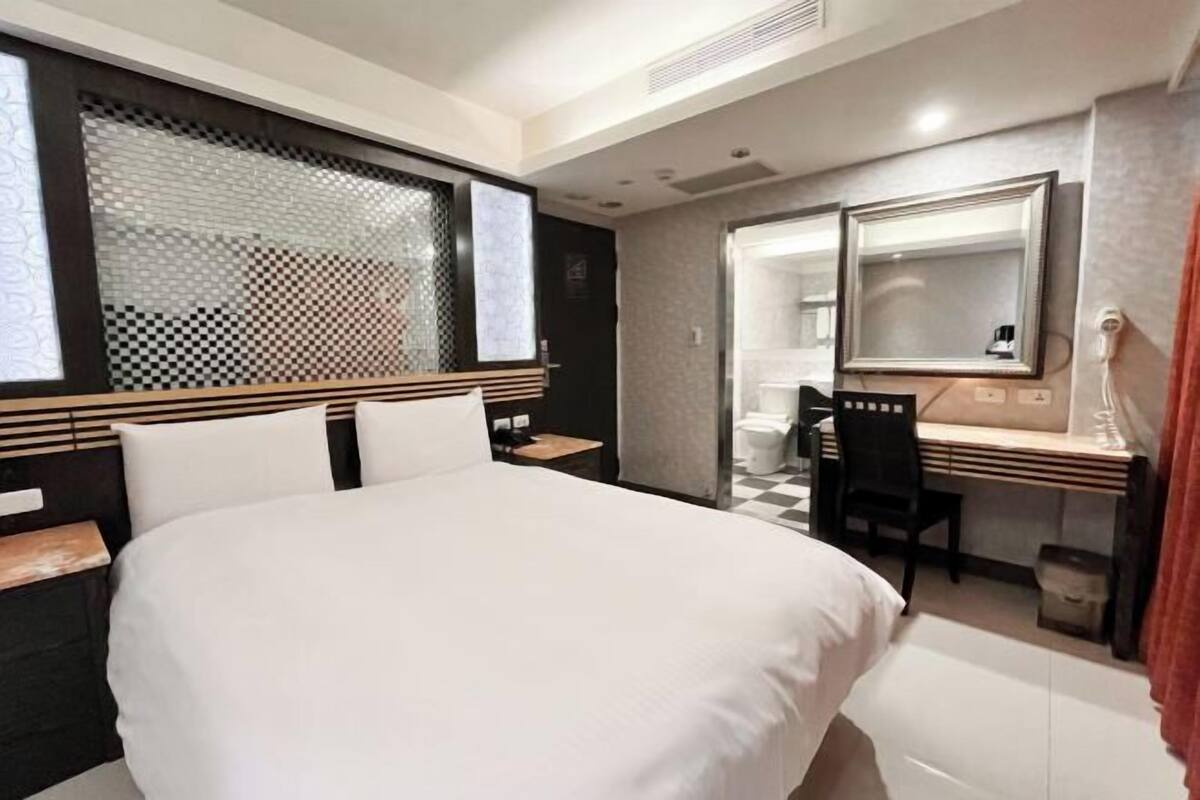 A hotel room with a bed, a desk, a chair, a mirror, and a window with a checkered pattern.