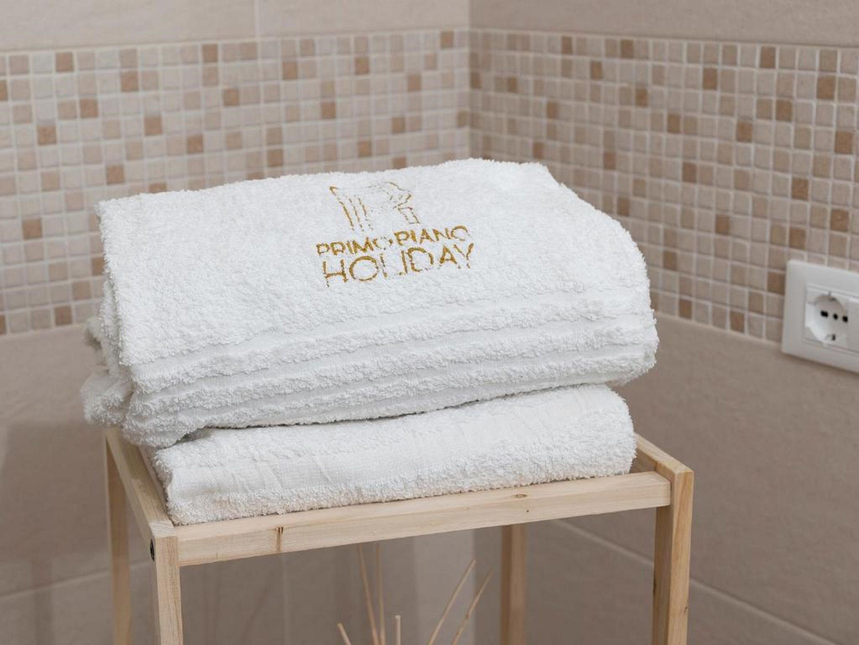 Una pila de toallas blancas con el bordado "PRINCIPAL HOLIDAY", sobre un taburete de madera en un baño con paredes de azulejos color beige.
