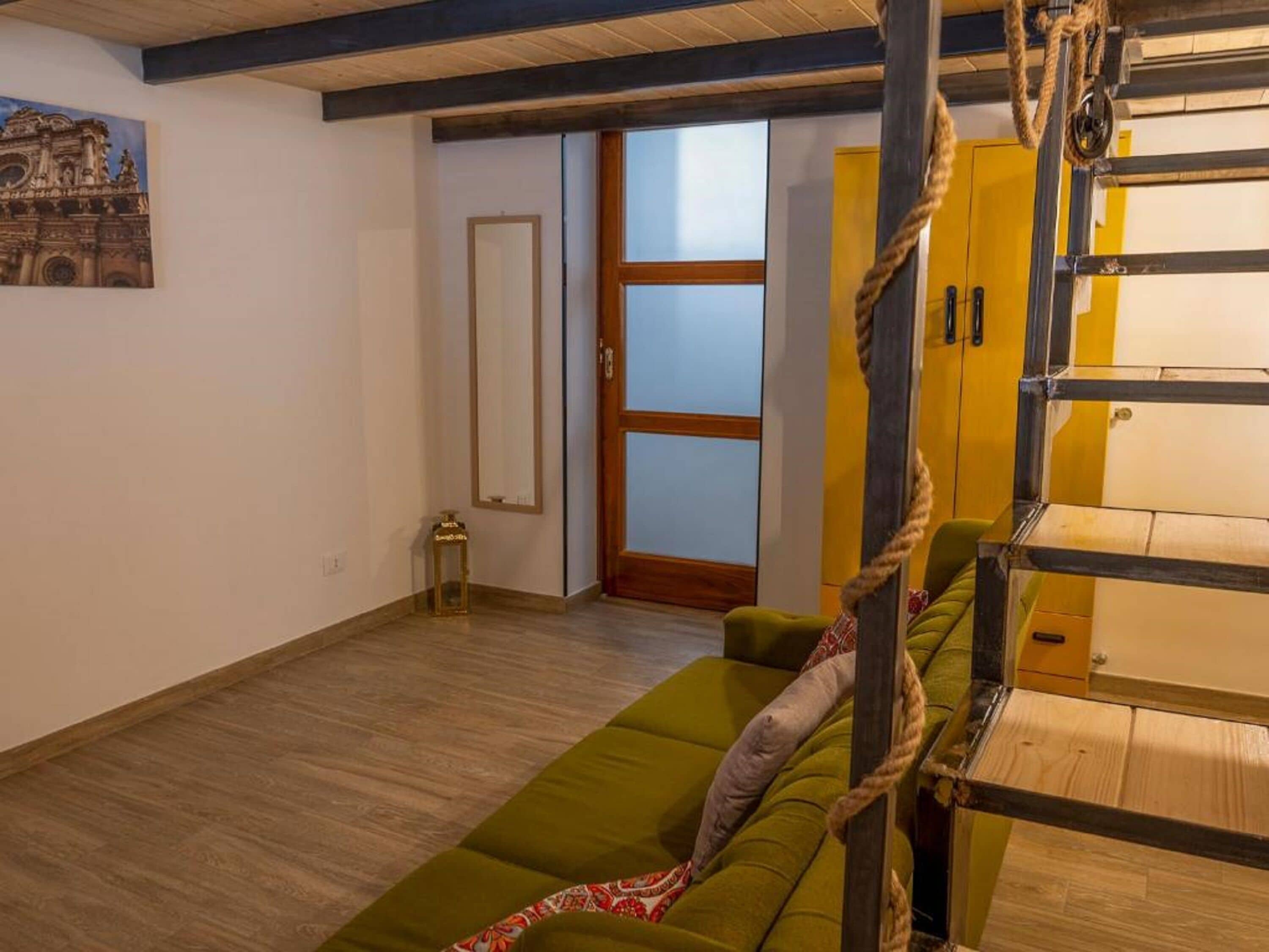 Una habitación con un sofá verde, piso de madera, una escalera de cuerda y una puerta con paneles de vidrio esmerilado.