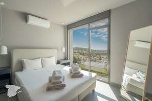 A modern bedroom with a large bed, a view of a cityscape through a sliding glass door, and a mirror on the wall. - CostaBlancaDreams - Perla in Calpe (Calpe)