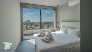 Une chambre d’hôtel moderne avec un grand lit, deux oreillers, une table de chevet et une vue sur le paysage urbain grâce à de grandes fenêtres du sol au plafond.
