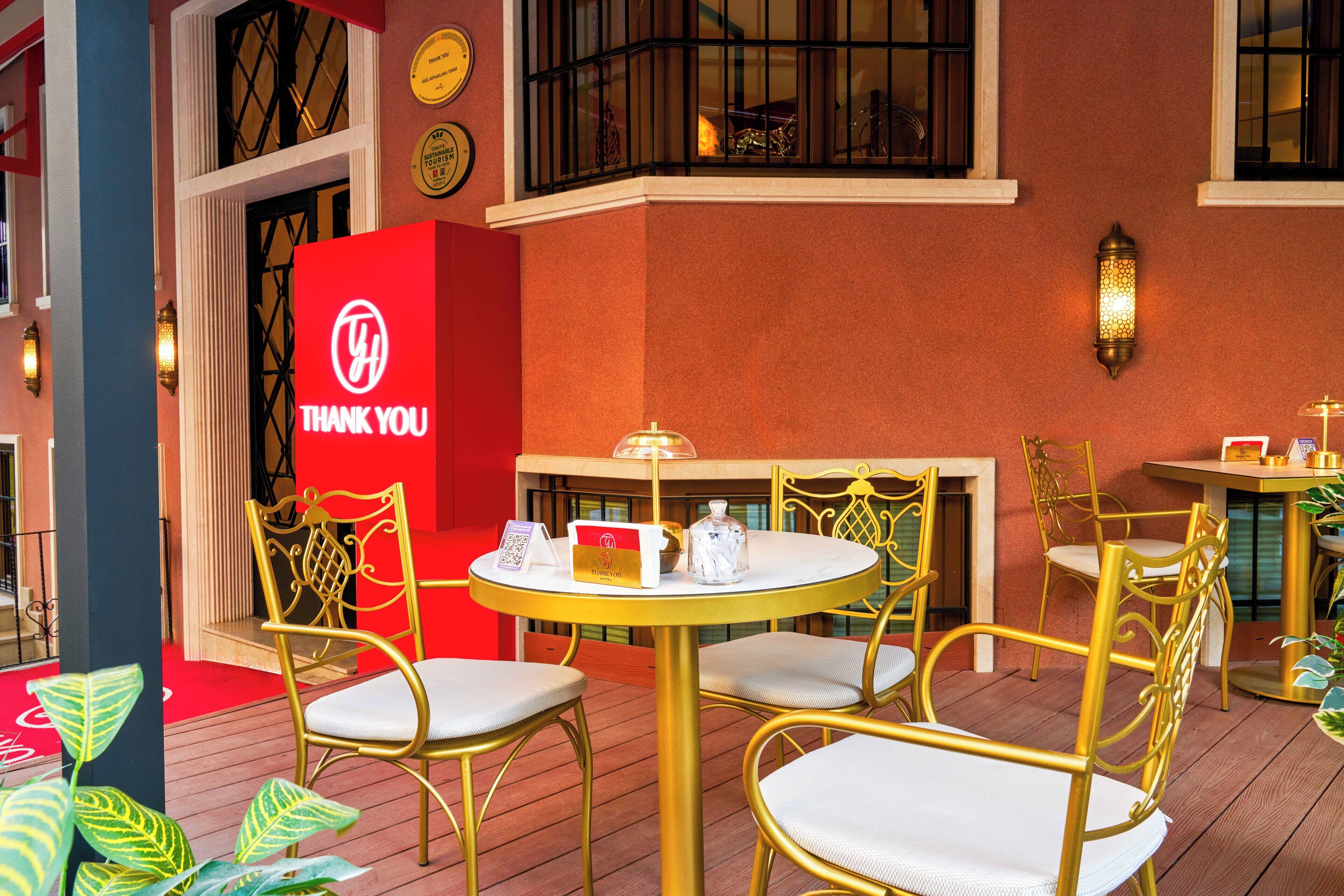 A terrace with round tables and gold chairs, a red 'THANK YOU' sign, and a decorative wall with a lantern.