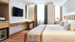 Ein Hotelzimmer mit einem großen Bett, einem flachbildfernseher an der Wand, einer Minibar und einem großen Fenster mit Vorhängen.