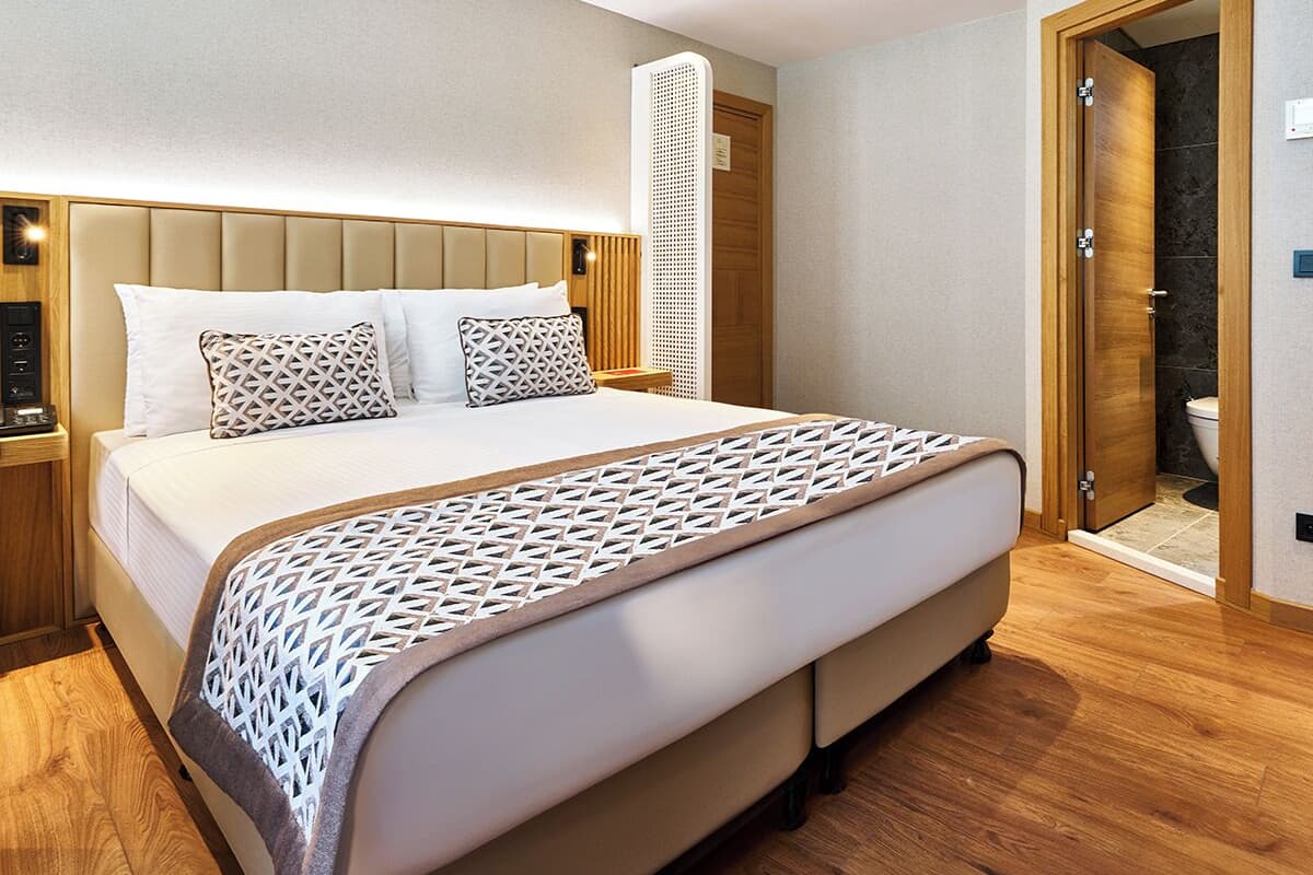 A modern hotel room with a large bed, a wooden headboard, a bedside table, and a bathroom visible through an open door.