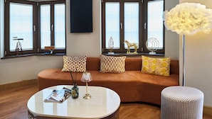 Ein modernes Wohnzimmer mit einer gebogenen Couch, einem runden Couchtisch und einem Beistelltisch. Auf der Couch liegen dekorative Kissen, eine Stehlampe steht bereit und ein Dekoelement befindet sich auf der Fensterbank.
