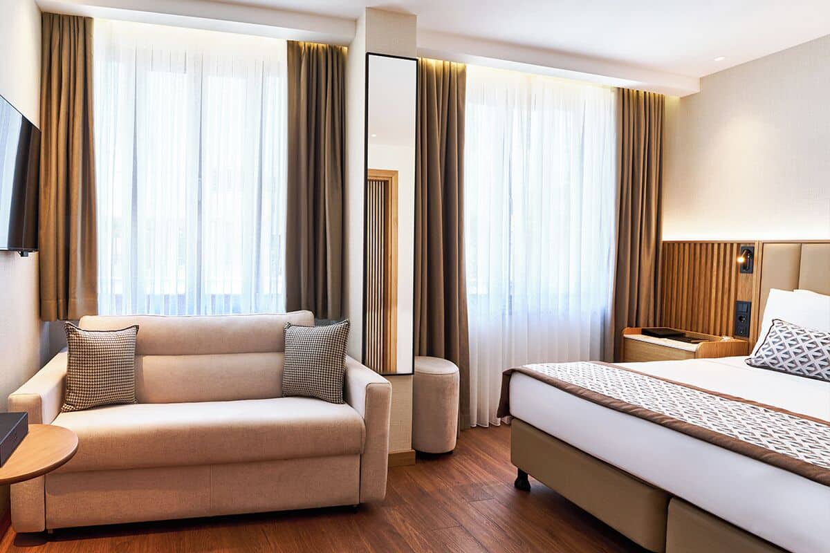 A modern hotel room with a sofa, a bed, a bedside table, and a TV.