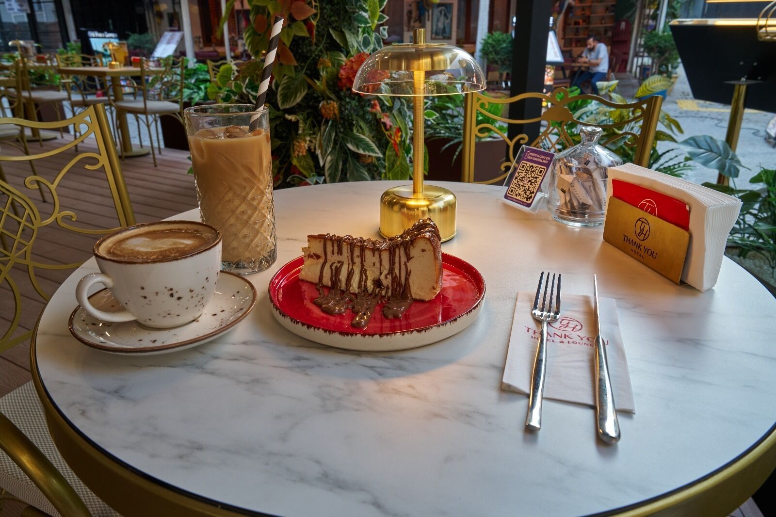 A table with a cup of coffee, a glass of iced coffee, a slice of cake with chocolate drizzle, a candle, a bottle of water, and a thank you card.
