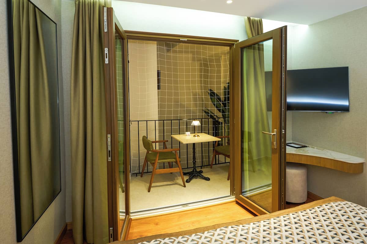 A hotel room with a balcony, a small table with a lamp, a TV mounted on the wall, and a patterned bedspread.