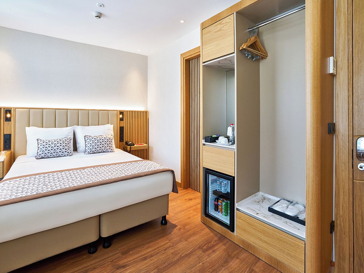A modern hotel room with a bed, a wardrobe with a built-in mini-fridge, and a small bedside table.