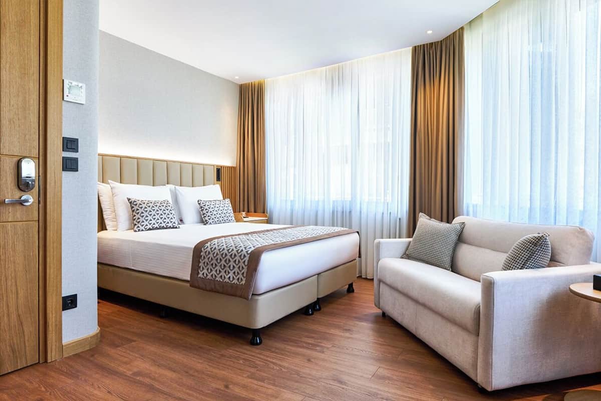A hotel room with a bed, a sofa, and a small table. There is a wooden floor and a window with curtains.
