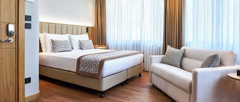 A hotel room with a bed, a sofa, and a small table. There is a wooden floor and a window with curtains.