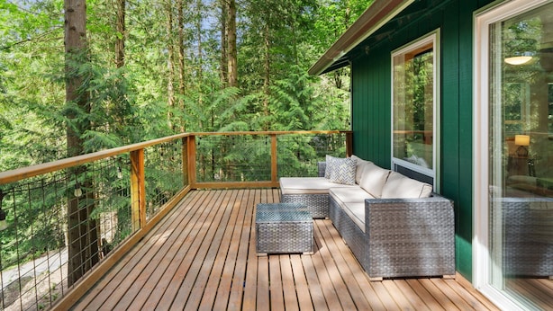 Eine Holzterrasse mit Glasgeländer, Korbmöbeln und Blick auf einen dichten Wald.