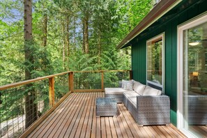 A wooden deck with a glass balustrade, wicker furniture, and a view of a dense forest. - Evergreen Cabin- serene getaway close to mountain biking, hiking & skiing (Bellingham)