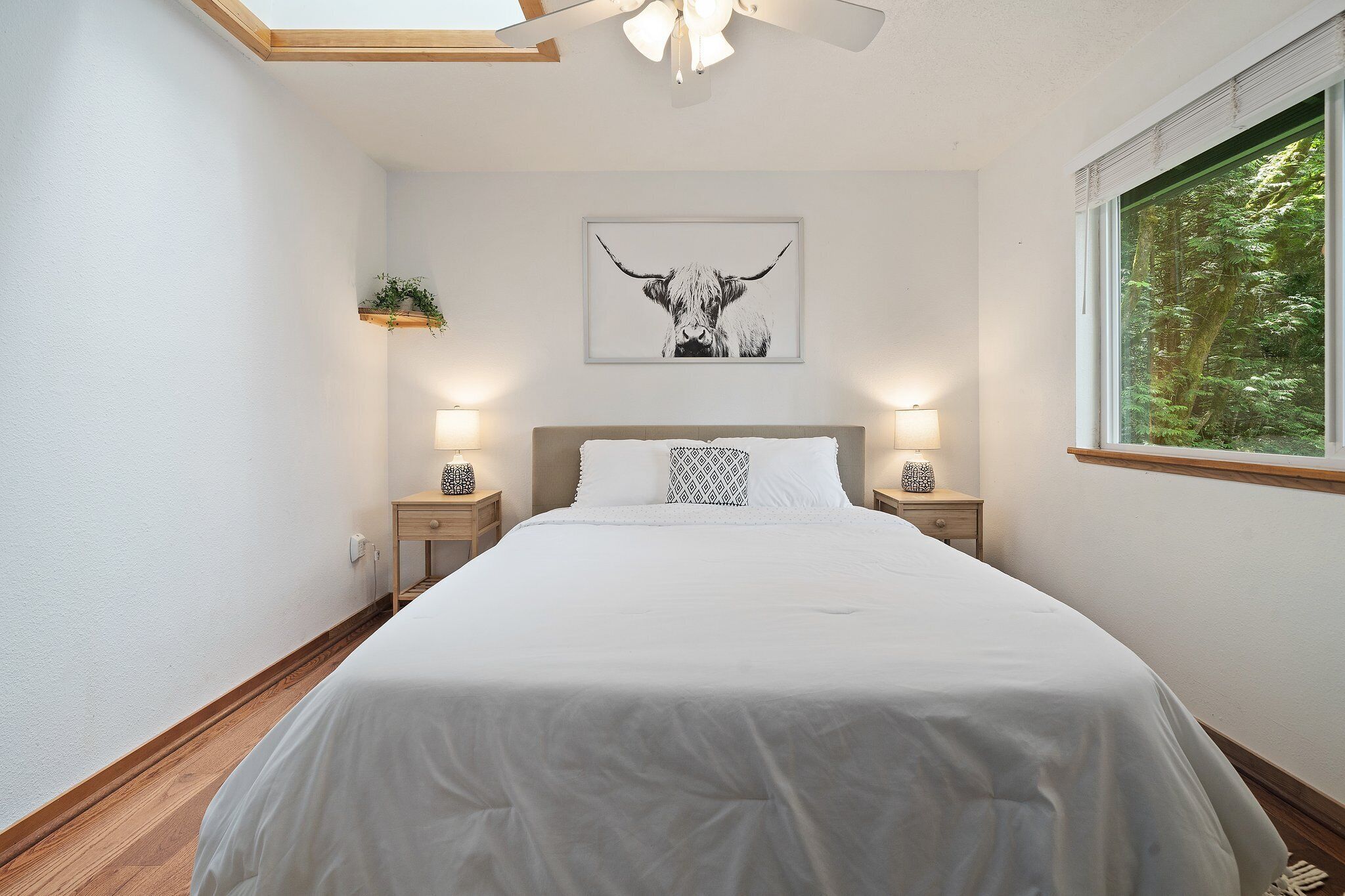 Une chambre avec un grand lit, deux tables de chevet, un ventilateur de plafond, une fenêtre donnant sur les arbres et une œuvre d’art encadrée accrochée au mur.