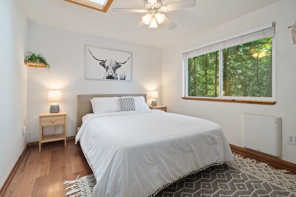 A bedroom with a bed, bedside lamps, a ceiling fan, a window with a view of trees, and a wall-mounted radiator. - Evergreen Cabin- serene getaway close to mountain biking, hiking & skiing (Bellingham)