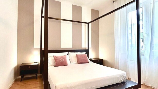 Un lit à baldaquin avec un matelas blanc et des oreillers roses, un plancher en bois et une table de chevet avec une lampe.