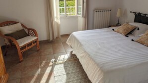 Una camera da letto con un letto, una sedia, una finestra con tende e termosifone, con vista sul verde.