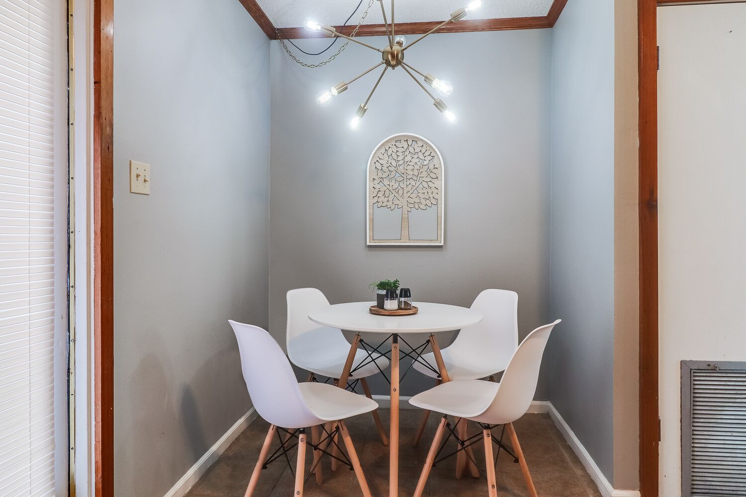 A small dining area with a round table, four white chairs, a chandelier, and a wall-mounted tree artwork.