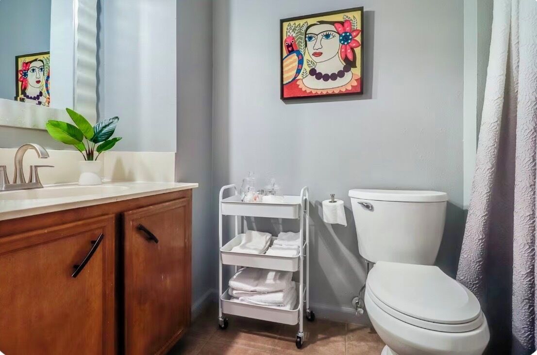 Un baño con un lavamanos blanco, armarios de madera, un inodoro blanco, un carrito de toallas blanco y un cuadro en la pared.