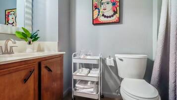 Un baño con un lavamanos blanco, armarios de madera, un inodoro blanco, un carrito de toallas blanco y un cuadro en la pared.