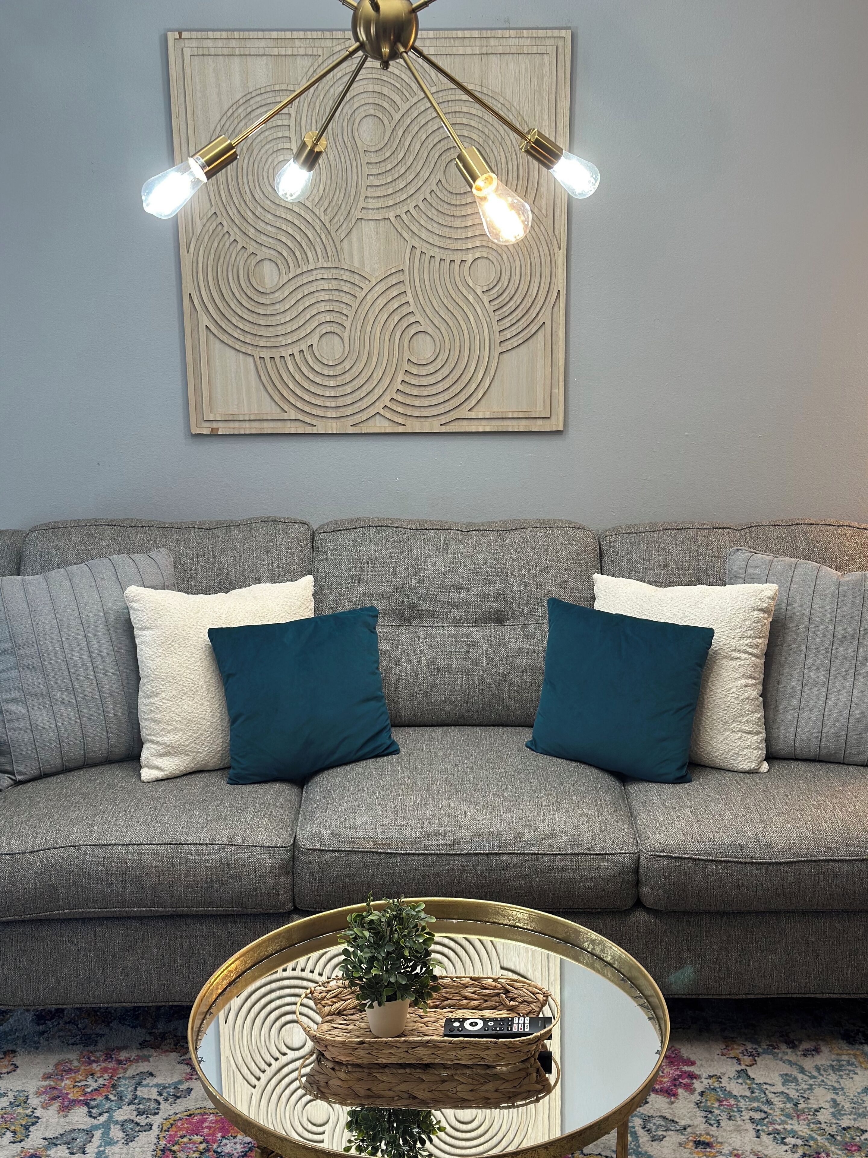 A living room with a grey sofa, two blue and two white cushions, a round coffee table with a plant and remote control, and a wall-mounted light fixture.