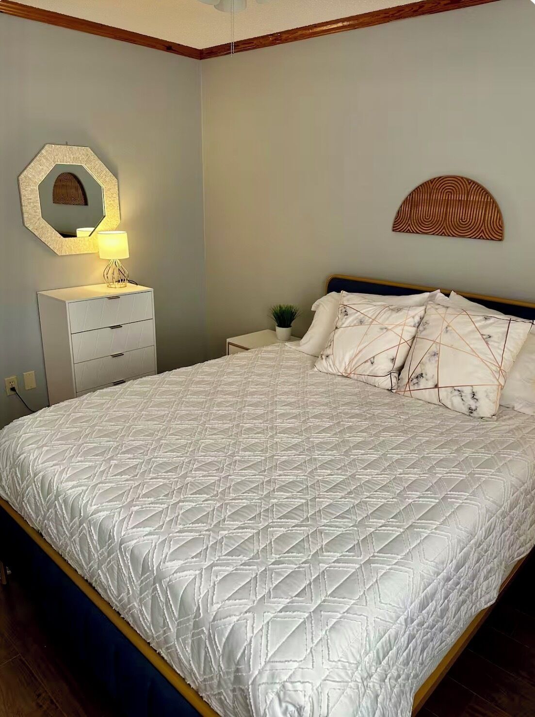 Un lit bien fait, recouvert d’une courtepointe blanche, une table de chevet avec une lampe et un miroir fixé au mur.